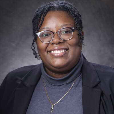A smiling Black woman with braided hair, wearing glasses, a gray turtleneck, and a black blazer.