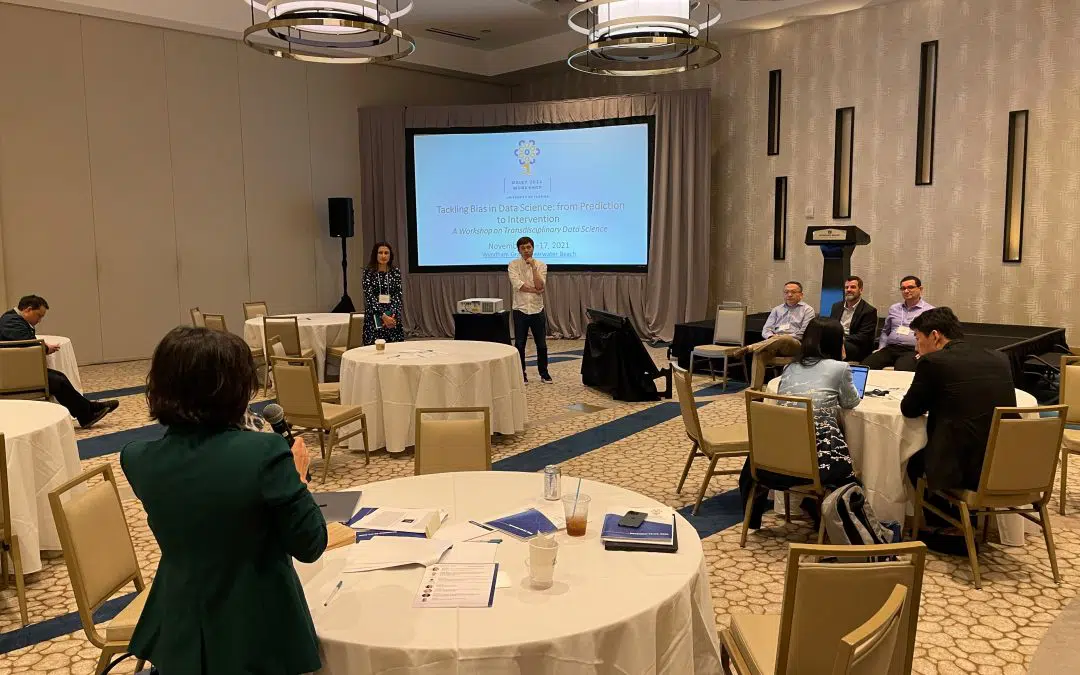 Conference room with a large presentation screen. A group of people ask questions to two presenters of a research presentation.