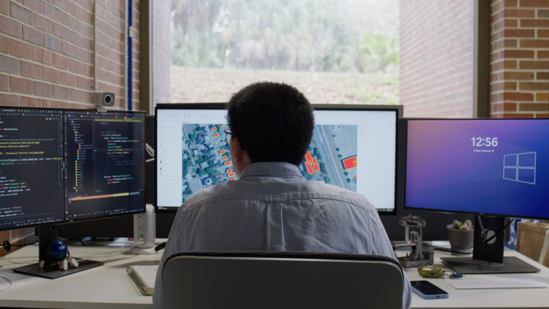 A man sitting in front of a computer monitor while facing a window.