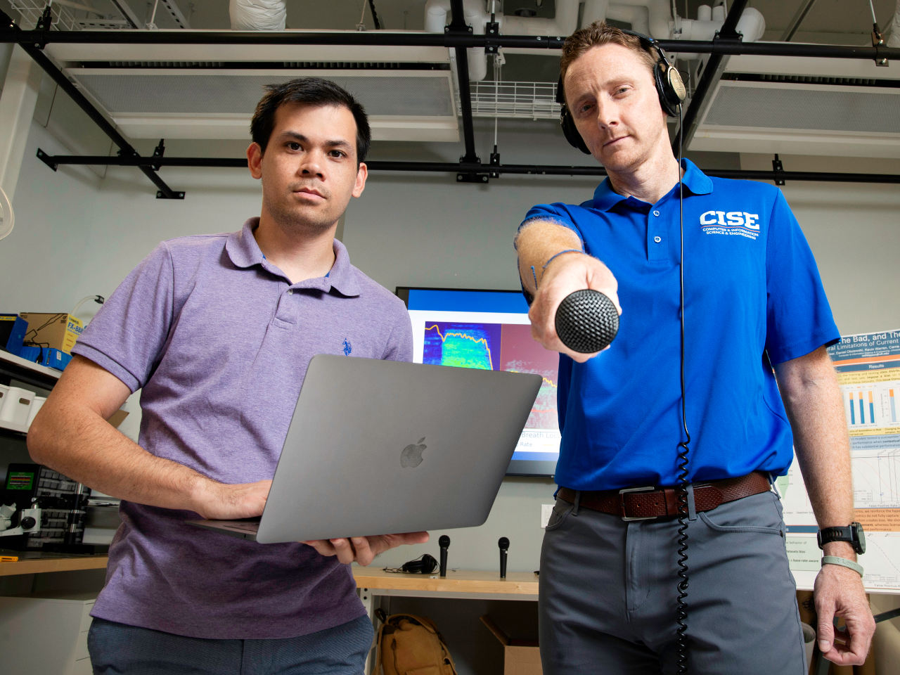 Two men standing facing the camera, one holding a laptop and the other wearing headphones and holding a microphone at the camera